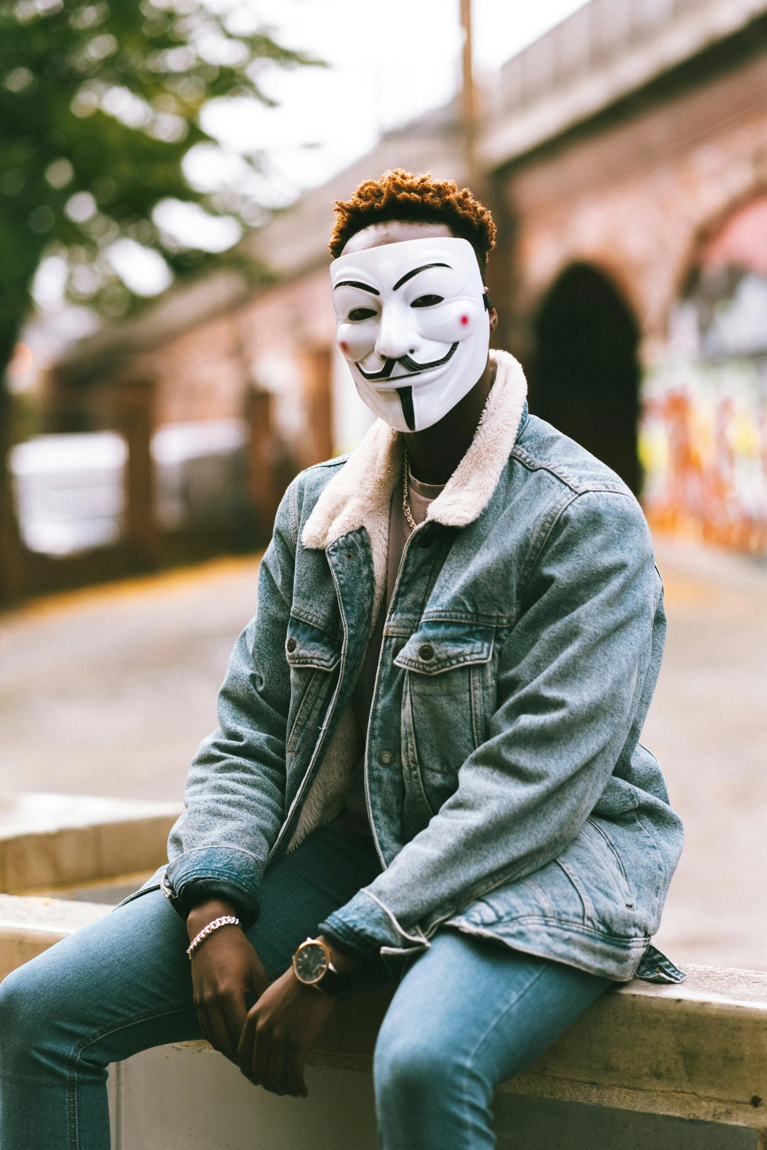 a person wearing a mask sitting on a bench, a photo, trending on pexels, graffiti, attractive androgynous humanoid, anonymous lion face, wearing a jeans jackets, portrait of professor sinister