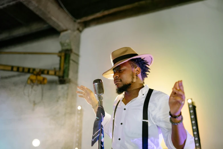 a man in a hat and suspenders holding a microphone, by Chinwe Chukwuogo-Roy, pexels, happening, square, soft light, artisanal art, avatar image