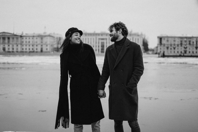 a black and white photo of a couple holding hands, by Emma Andijewska, pexels, antipodeans, wearing a long coat, smiling couple, saint petersburg, on beach