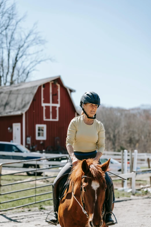 a woman riding on the back of a brown horse, a portrait, by Alison Geissler, trending on unsplash, red barn in distance, wearing a helmet, older woman, gambrel roof