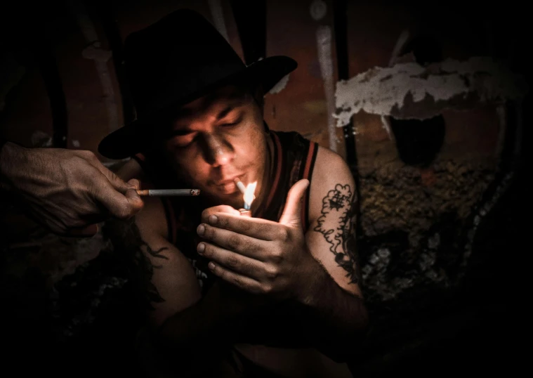 a man in a hat lighting a cigarette, a portrait, pexels contest winner, tattoo artist, murky dusty deep, pipe jungle, lit from below