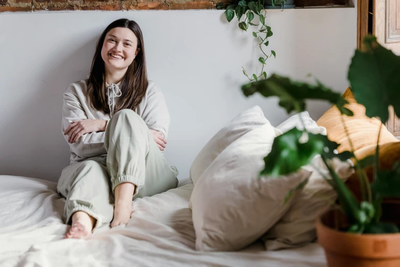 a woman sitting on top of a bed next to a potted plant, by Emma Andijewska, pexels contest winner, happening, wide grin, wearing pajamas, wearing a linen shirt, charli bowater and artgeem