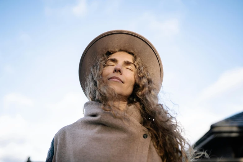 a woman with long hair wearing a hat, trending on pexels, looking up onto the sky, brown curly hair, brown cloak, portrait image