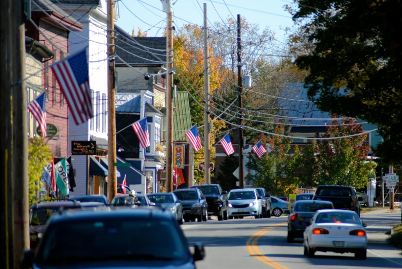 a street filled with lots of cars driving down it, a portrait, flickr, new hampshire, american flags, french village exterior, profile image