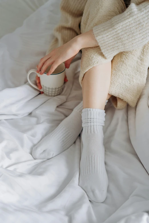 a woman sitting on a bed with a cup of coffee, inspired by Elsa Bleda, pexels contest winner, renaissance, white grey color palette, socks, all white, pale skin