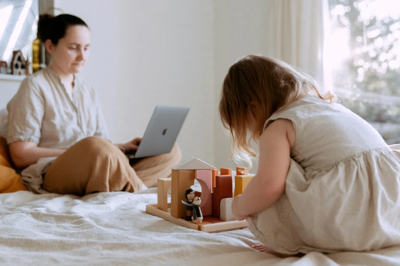 a woman sitting on top of a bed next to a little girl, pexels contest winner, working on her laptop, wooden art toys on base, healthcare, 1 4 9 3