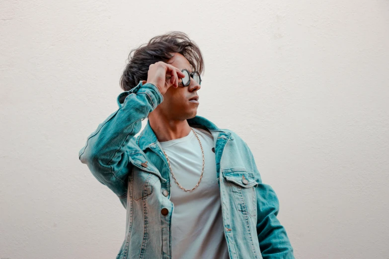 a man standing in front of a white wall talking on a cell phone, an album cover, by Robbie Trevino, trending on pexels, jean jacket, wear ray - ban glass, teal aesthetic, attractive pose