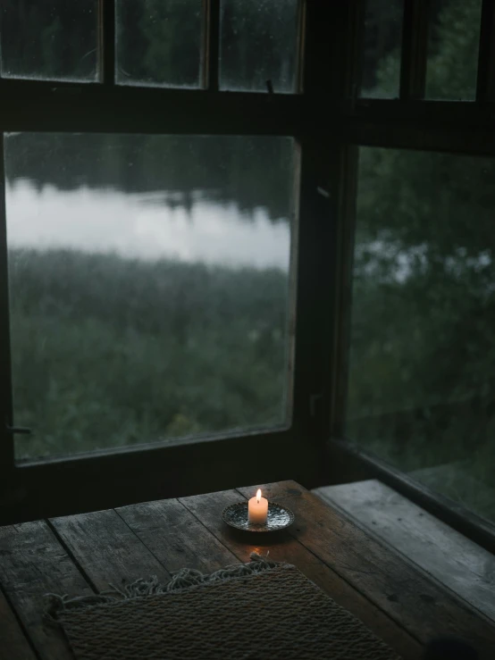 a candle sitting on top of a wooden table next to a window, a picture, trending on unsplash, on a lake, rainy window, light inside the hut, overcast dusk