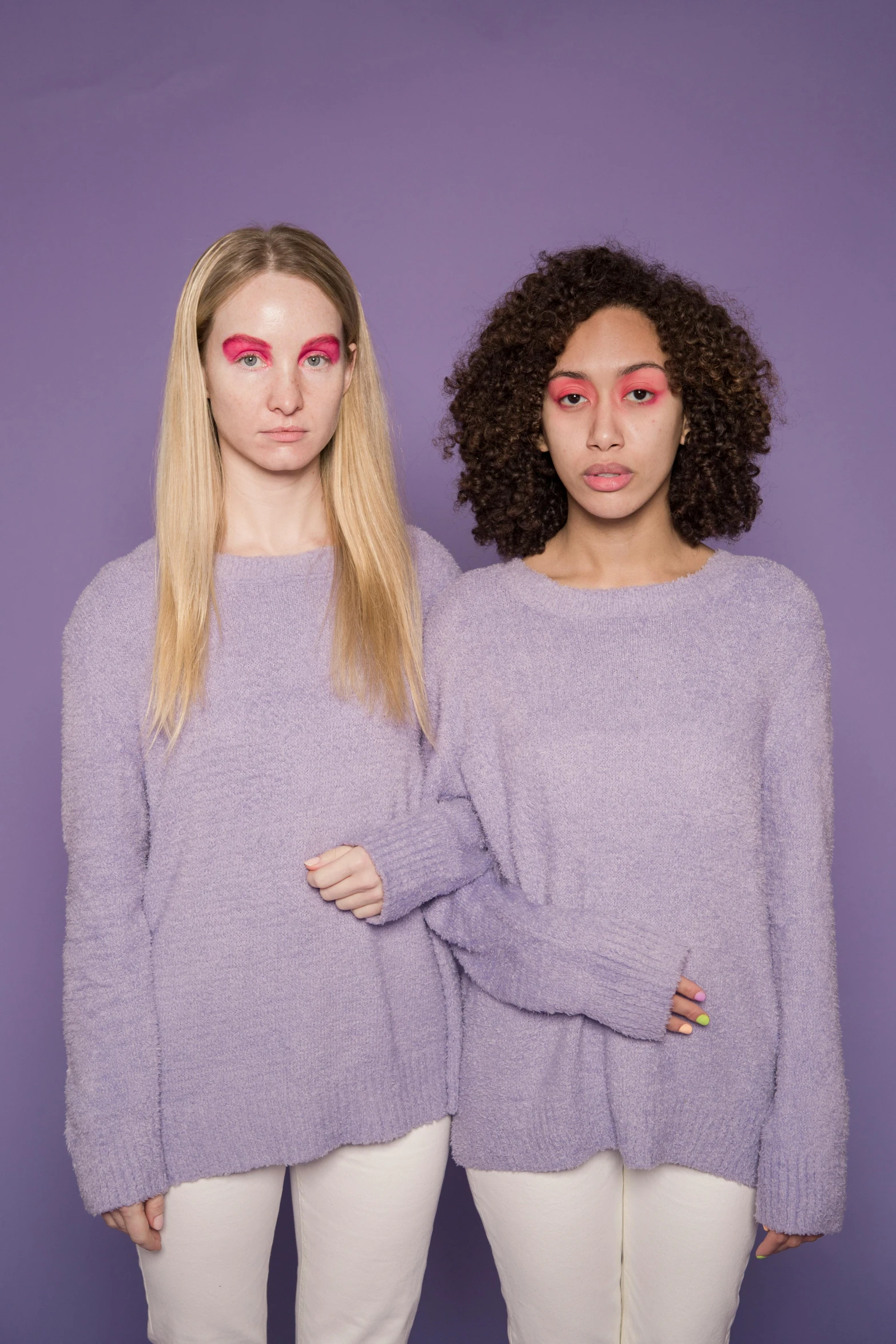 two women standing next to each other in front of a purple background, an album cover, inspired by Maud Naftel, trending on pexels, antipodeans, wearing a sweater, purple eye color, false human features, promotional picture