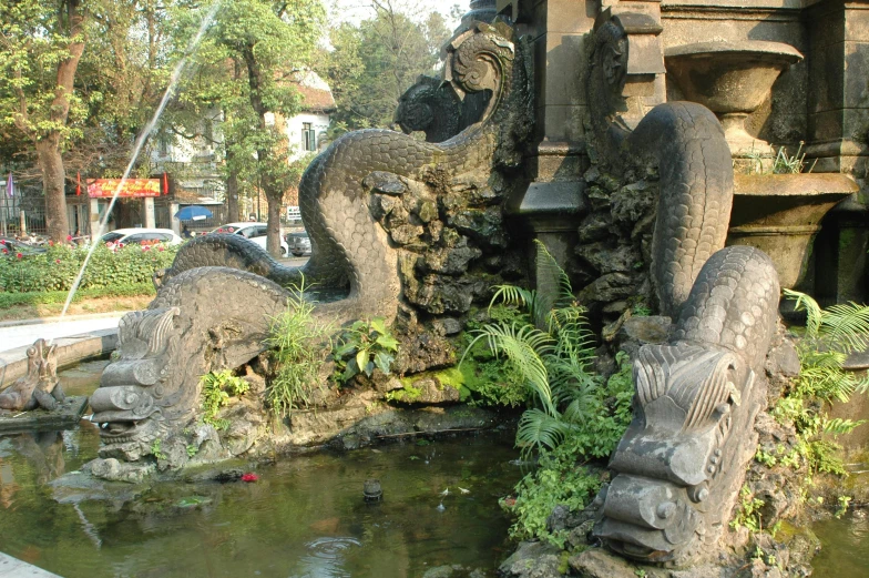 a group of statues sitting next to a body of water, a statue, dragon tail, city of the jungle, lush surroundings, street corner