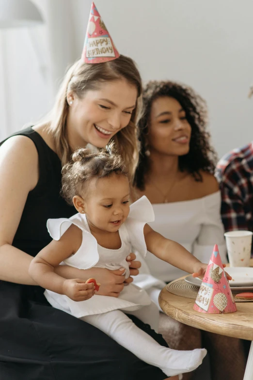 a group of people sitting around a table with a cake, toys, with a kid, woman holding another woman, profile image