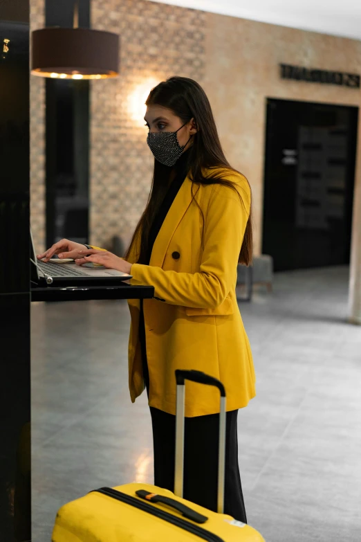 a woman in a yellow jacket is using a laptop, private press, wearing all black mempo mask, in a hotel hallway, standing on a desk, wearing nanotech honeycomb robe