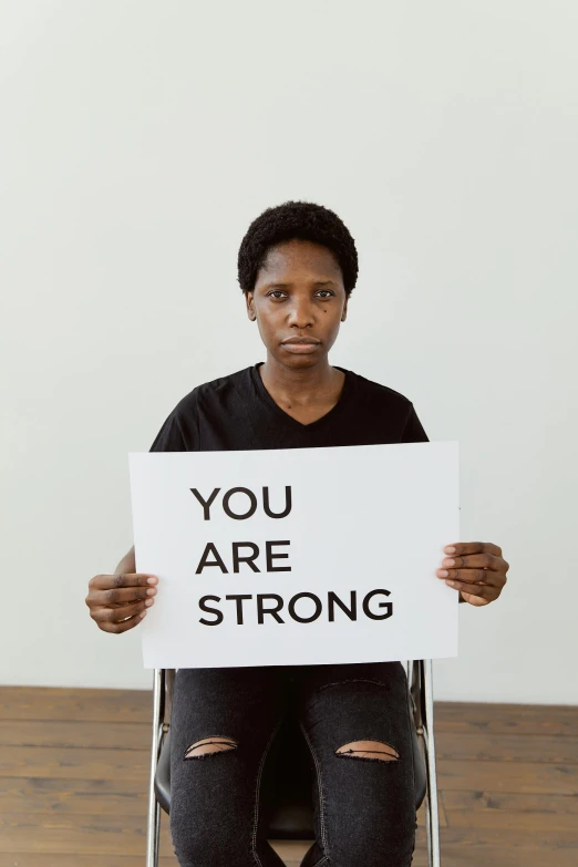 a woman sitting in a chair holding a sign that says you are strong, a black and white photo, standing together, dark-skinned, health supporter, with a white background