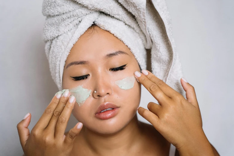 a woman with a towel on her head putting cream on her face, by Julia Pishtar, trending on pexels, pale green glow, an asian woman, square, manuka