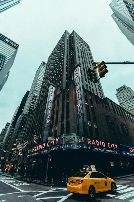 a yellow taxi driving down a street next to tall buildings, by Adam Rex, pexels contest winner, modernism, photo of a big theaterstage, new york buildings, buildings carved out of stone, concert