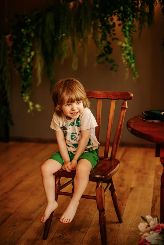 a little boy sitting on top of a wooden chair, next to a plant, green shirt, on a wooden table, dressed in a top and shorts