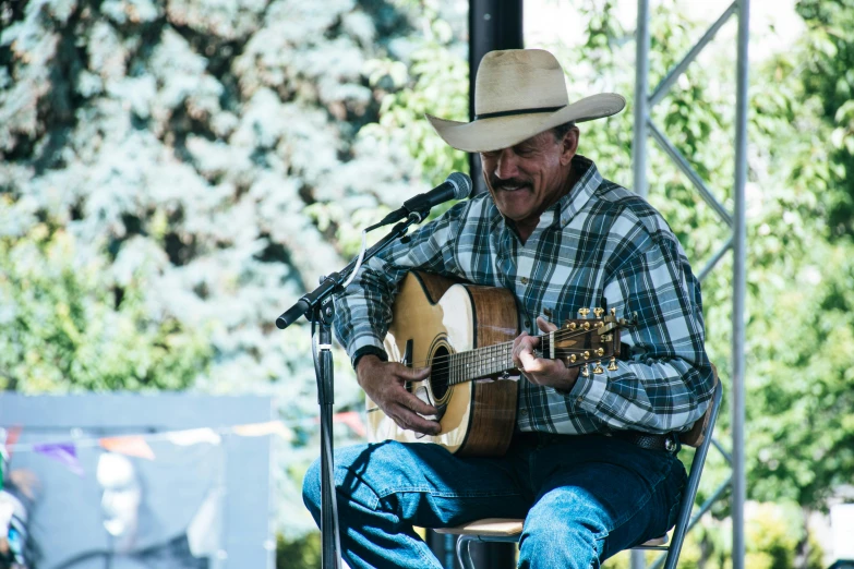 a man in a cowboy hat playing a guitar, by Jim Nelson, unsplash, square, festivals, idaho, sitting in front of a microphone