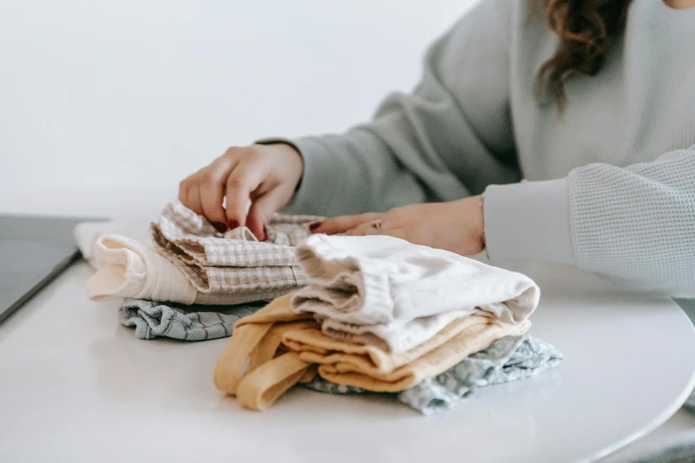 a woman sitting at a table with a pile of cloth, trending on unsplash, on a pale background, touching her clothes, earthy colours, stitching