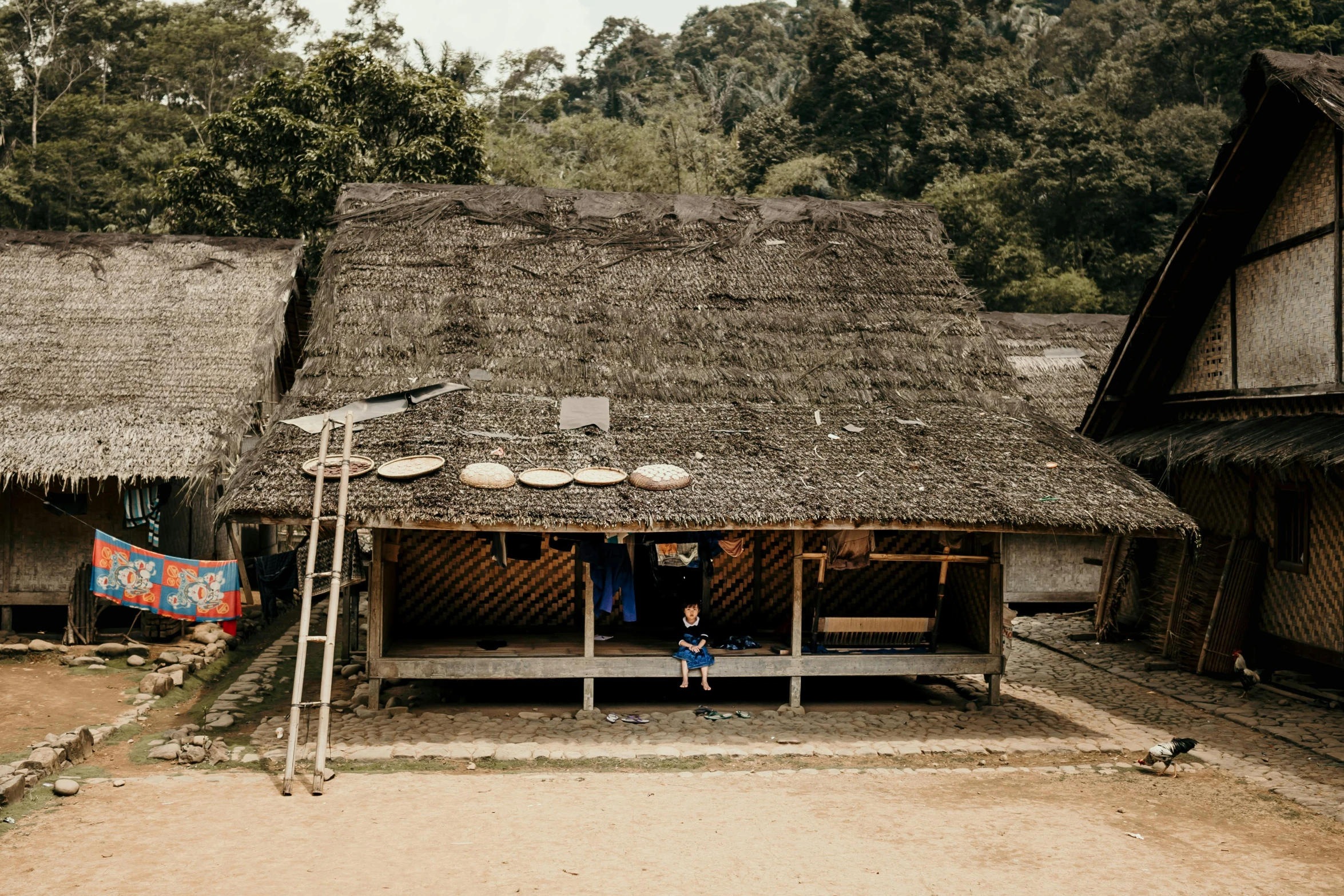 a person sitting on a bench in front of a hut, by Emma Andijewska, pexels contest winner, sumatraism, 4yr old, wide high angle view, standing outside a house, avatar image