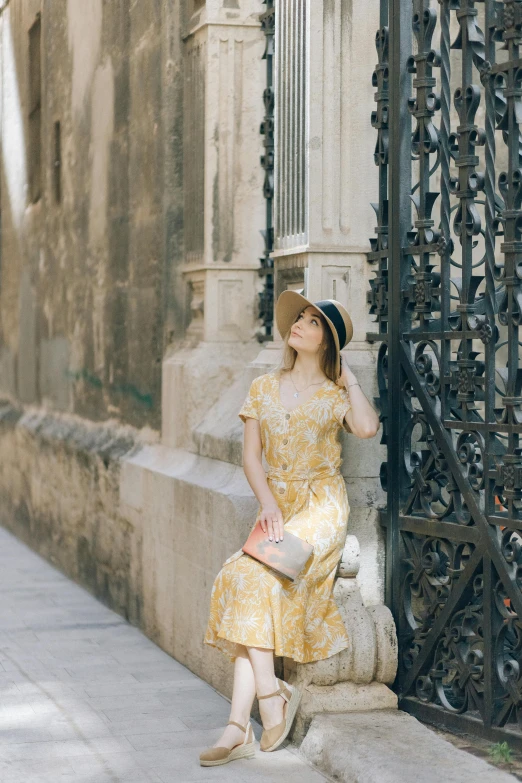 a woman in a yellow dress and hat sitting on a wall, inspired by Luis Paret y Alcazar, unsplash, baroque, leaning on door, square, delicate patterned, low quality photo