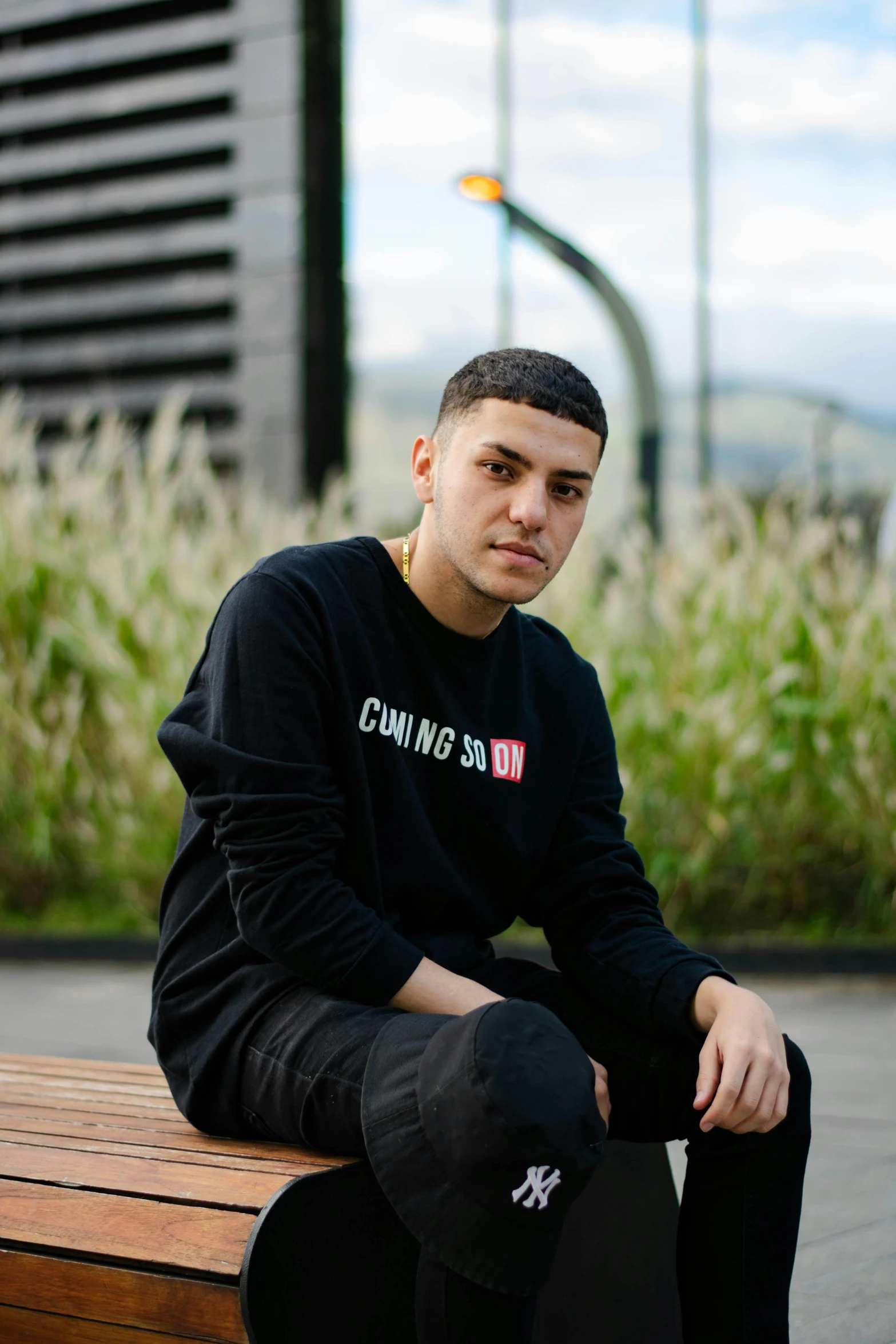 a man sitting on top of a wooden bench, an album cover, inspired by Carlos Berlanga, unsplash, wearing a black sweater, yzy gap, looking to camera, ismail