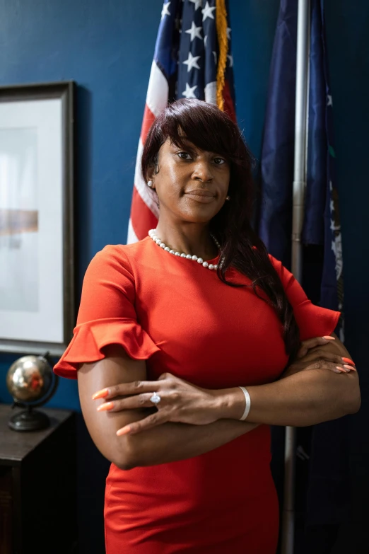 a woman in a red dress standing in front of an american flag, jemal shabazz, in the office, arms crossed, senator armstrong