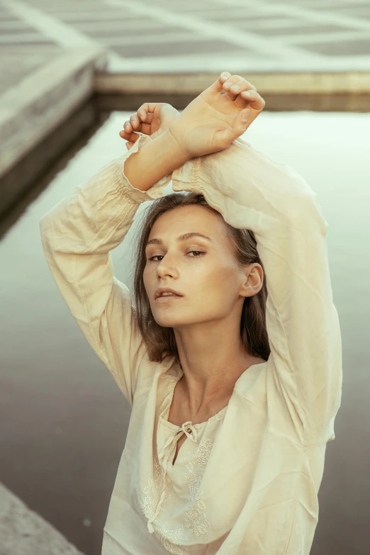 a woman standing next to a body of water, an album cover, inspired by Emma Andijewska, pexels contest winner, renaissance, cream colored peasant shirt, cheekbones, with a cool pose, sunfaded