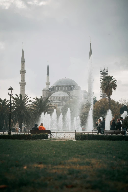 a group of people sitting on a bench in front of a fountain, inspired by Altoon Sultan, pexels contest winner, hurufiyya, minarets, trees in foreground, square, people walking around