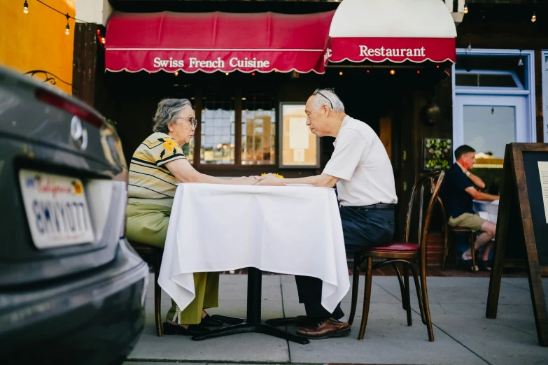 a couple of people sitting at a table in front of a restaurant, unsplash, 7 0 years old, irina french, two frail, sydney hanson