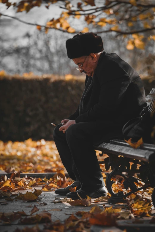 a man sitting on a bench looking at his cell phone, pexels contest winner, covered in fallen leaves, wearing black old dress and hat, man with glasses, orthodox