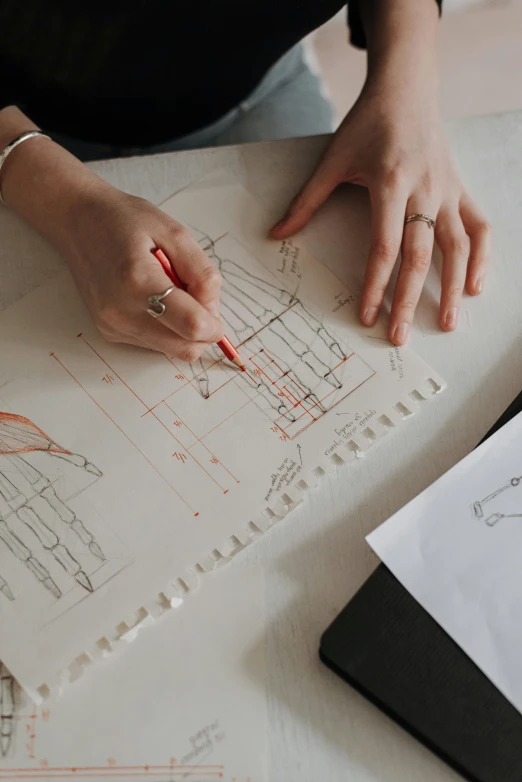 a woman sitting at a table drawing on a piece of paper, analytical art, blueprints, wearing a designer top, knolling, on vellum