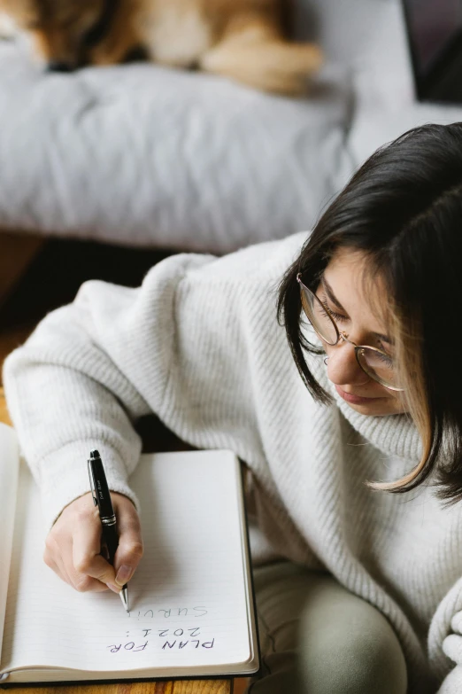 a woman sitting on a couch writing in a notebook, pexels contest winner, wearing a white sweater, wavy long black hair and glasses, thumbnail, someone sits in bed