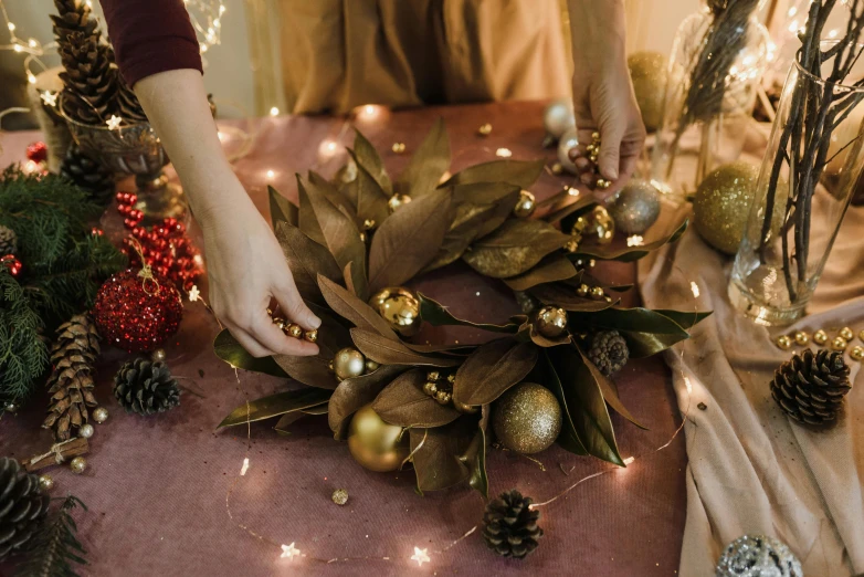 a close up of a person placing a wreath on a table, by Julia Pishtar, pexels contest winner, brown and gold, firefly lights, magnolia leaves and stems, crown made of fabric