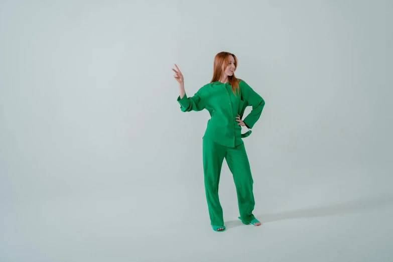 a woman in a green jumpsuit on a white background, by Marina Abramović, trending on pexels, fantastic realism, young redhead girl in motion, waving, wearing a colorful men's suit, standing in corner of room