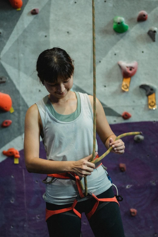 a woman holding a rope while standing in front of a climbing wall, ben lo, profile image, in a workshop, mid 2 0's female