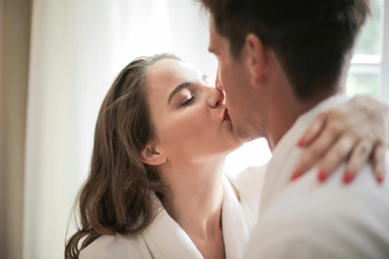 a man and a woman kissing in front of a window, pexels, wearing white silk robe, manuka, brunette, ad image