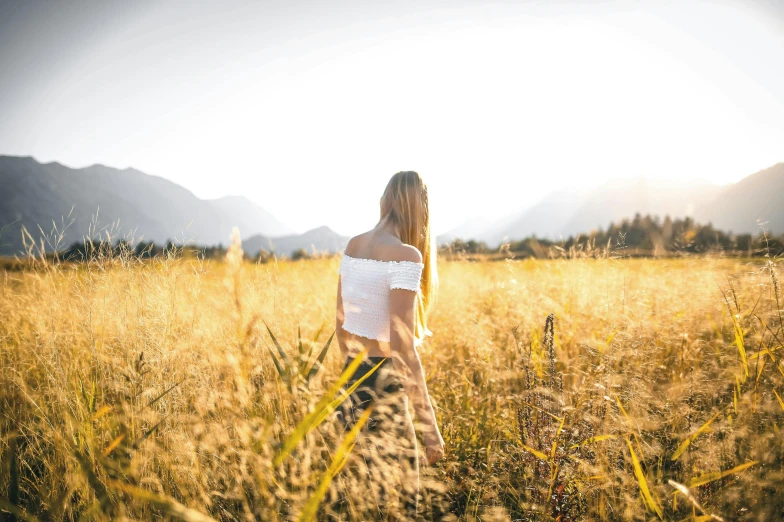 a woman standing in a field of tall grass, pexels contest winner, mountains in the background, golden hues, instagram post, walking away