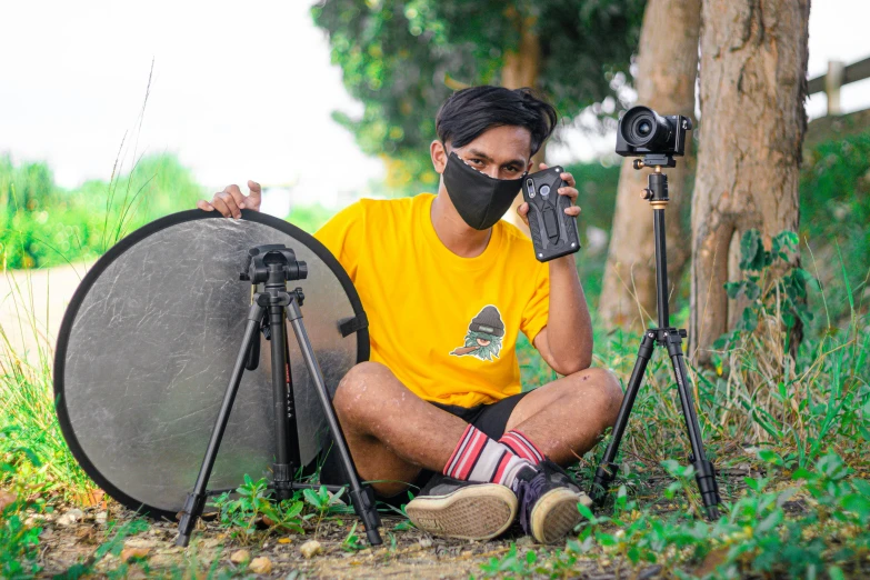 a man sitting on the ground next to a camera, avatar image, wearing yellow croptop, outdoor photo, tripod
