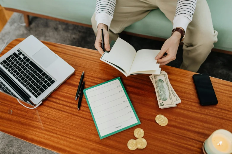 a person sitting on a couch with a laptop and money, pexels contest winner, private press, writing on a clipboard, standing on a desk, wooden desks with books, 🦩🪐🐞👩🏻🦳