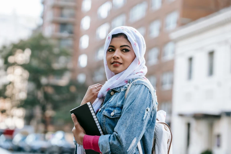 a woman in a hijab is holding a book, trending on unsplash, hurufiyya, standing in township street, proud smirk, at college, patterned scarf