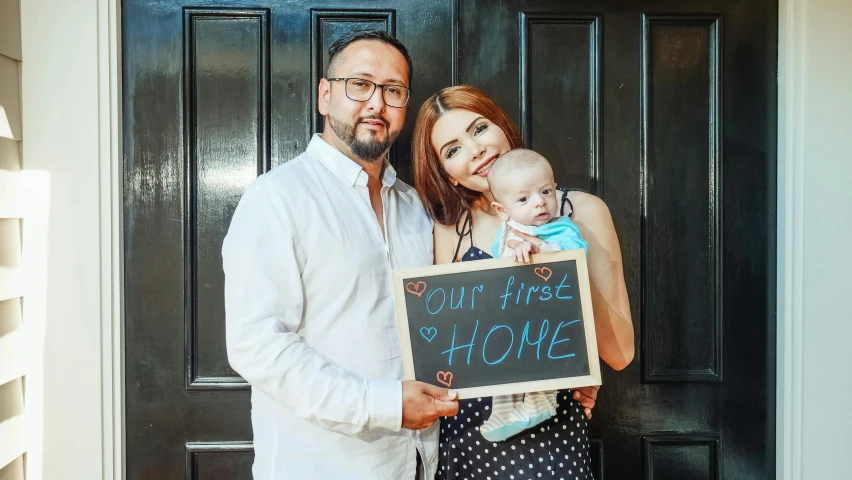 a man and woman holding a baby in front of a door, ginger bearded man with glasses, house and home, woman holding sign, portrait image