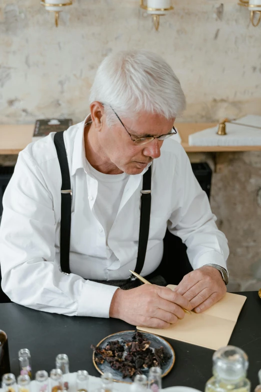 a man sitting at a table writing on a piece of paper, inspired by Karl Gerstner, pexels contest winner, john pawson, wearing an apron, gold and silver ink, older male