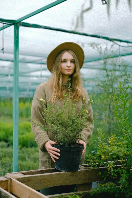 a woman holding a potted plant in a greenhouse, a picture, inspired by Elsa Bleda, unsplash, renaissance, she is wearing a hat, wearing a green sweater, cara delevingne, manuka