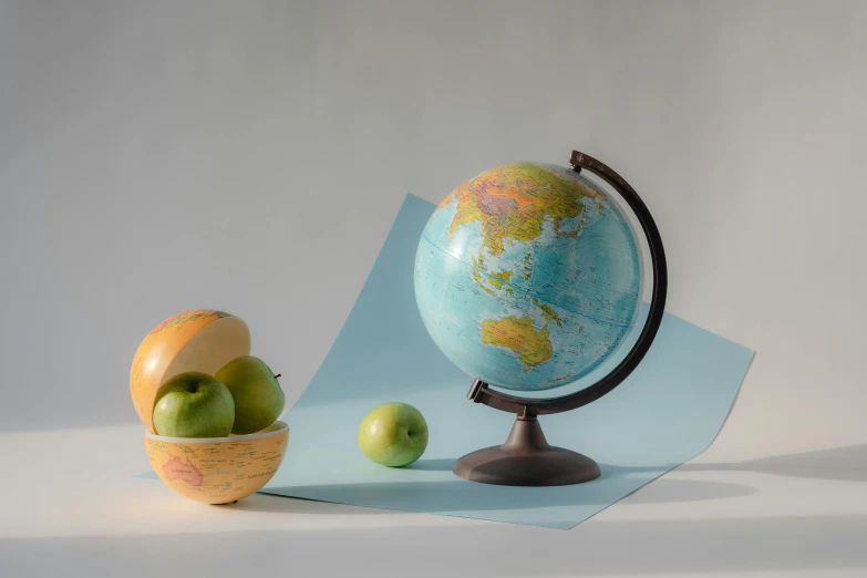 a globe sitting on top of a table next to apples, a still life, trending on unsplash, hyperrealism, paper modeling art, on a gray background, earth and pastel colors, half turned around