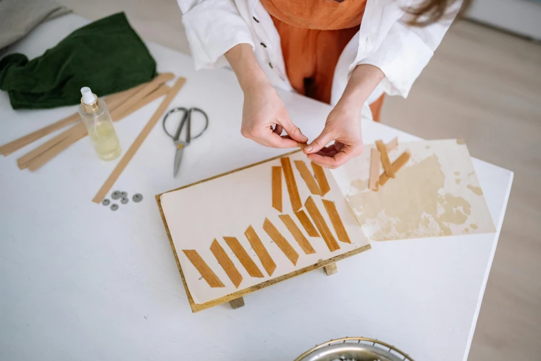 a woman in an orange apron working on a piece of art, by Emma Andijewska, trending on pexels, arts and crafts movement, cigarrette boxes at the table, inlaid with gold, on white background, made of woods