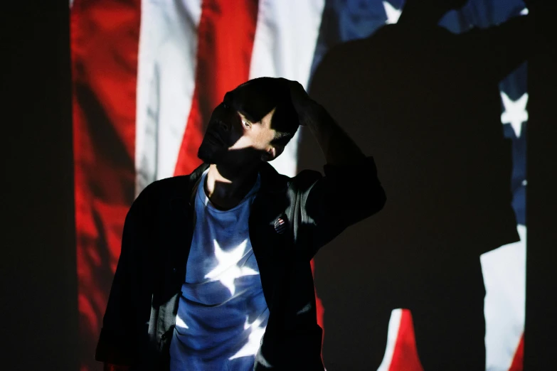 a man standing in front of an american flag, an album cover, by Attila Meszlenyi, unsplash, hyperrealism, finn wolfhard, projection mapping, facepalm, performing