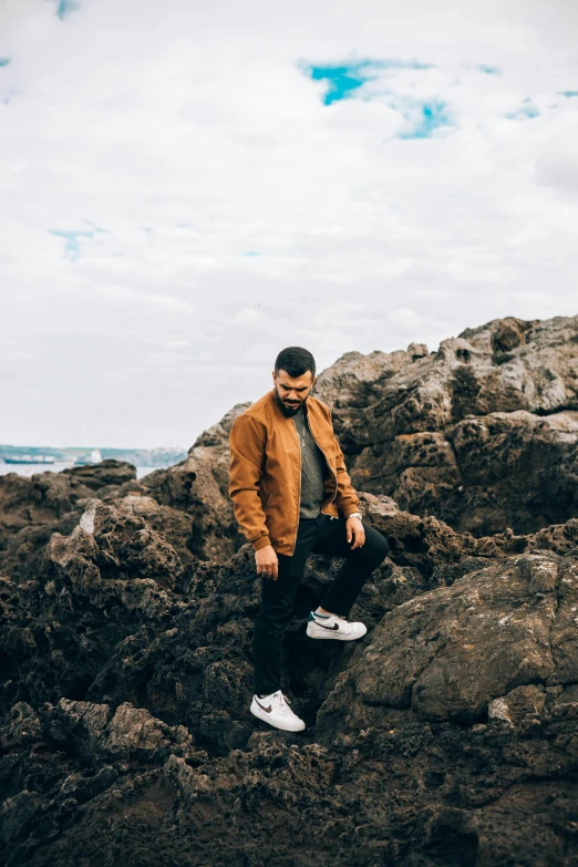 a man sitting on a rock near the ocean, an album cover, pexels contest winner, happening, wearing a bomber jacket, standing on boulder, caramel. rugged, trending on r/techwearclothing