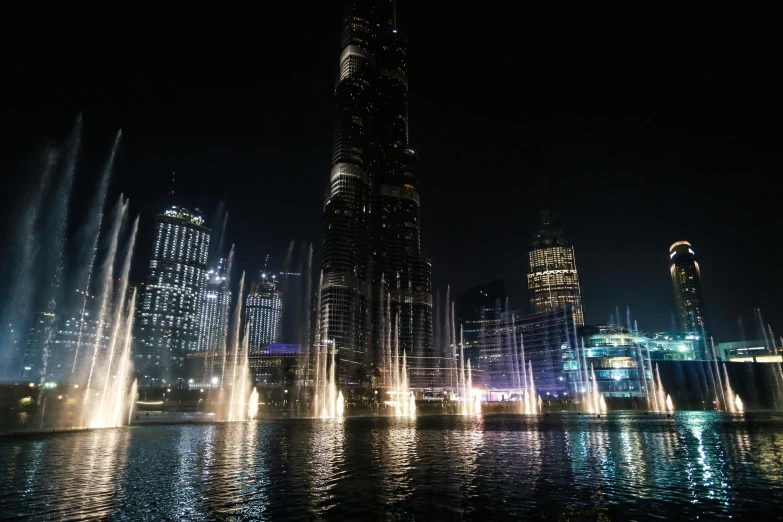 a water fountain in the middle of a city at night, a detailed matte painting, pexels contest winner, hurufiyya, gta : dubai, tall metal towers, hd footage, thumbnail