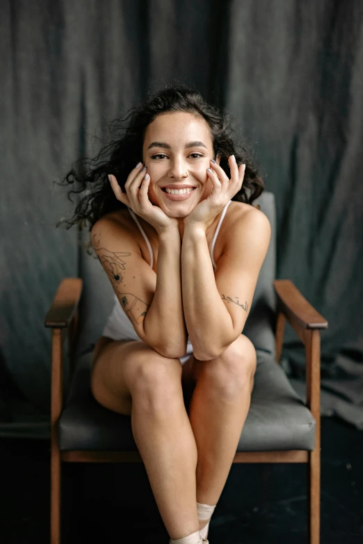 a beautiful young woman sitting on top of a chair, pexels contest winner, antipodeans, dark short curly hair smiling, hands behind her body pose!, oona chaplin, wearing a camisole and shorts