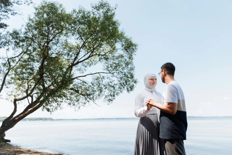 a man and a woman standing next to a body of water, by Julia Pishtar, hurufiyya, reaching out to each other, trees in background, riyahd cassiem, on a bright day